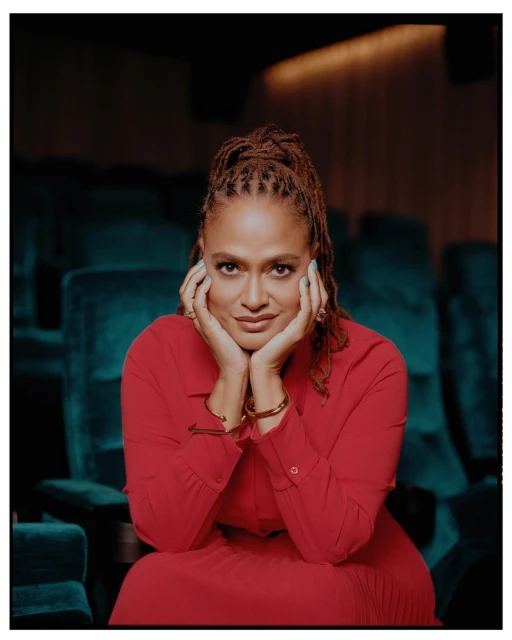 A woman with braided hair, wearing a red dress, is seated in a theatre with her hands near her face, smiling in a medium shot.