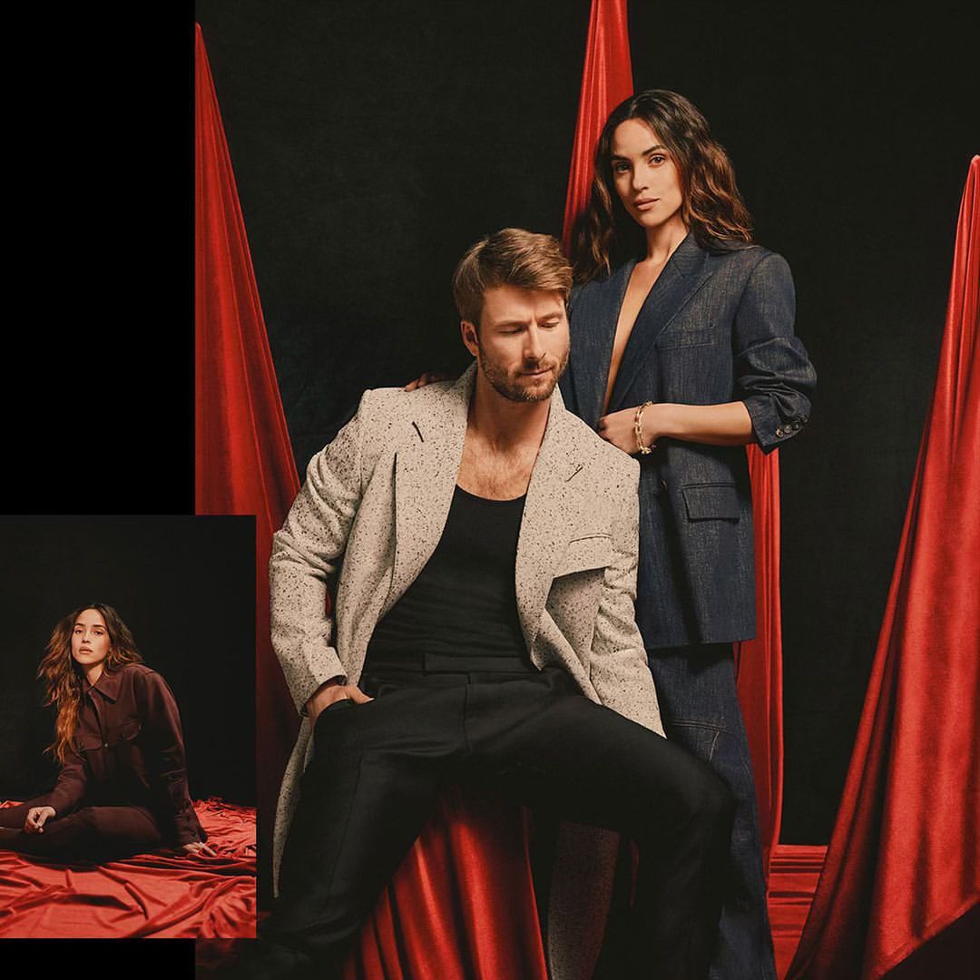 Man and woman posing in studio with red fabric drapes. The man is seated wearing a light-colored coat and dark pants. She wears a denim suit.