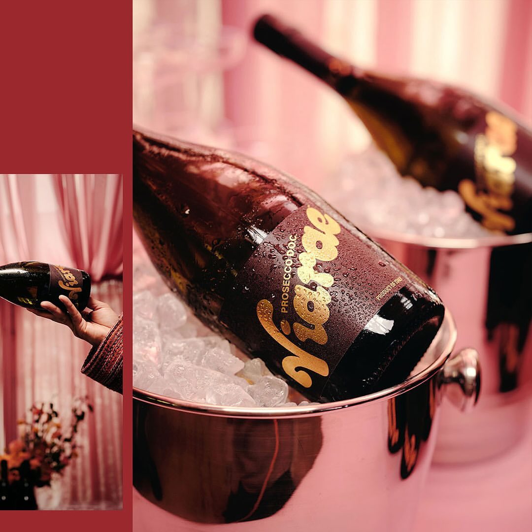 Two bottles of prosecco in ice-filled buckets, one held by a hand, set against a pink backdrop with flowers. Celebratory drink.