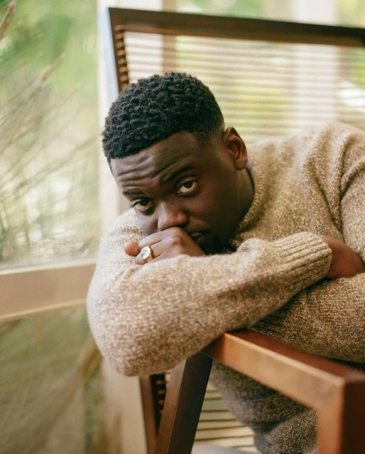 Close-up portrait of an African American man with short dark hair sitting in a wooden chair near a window with natural lighting indoors.