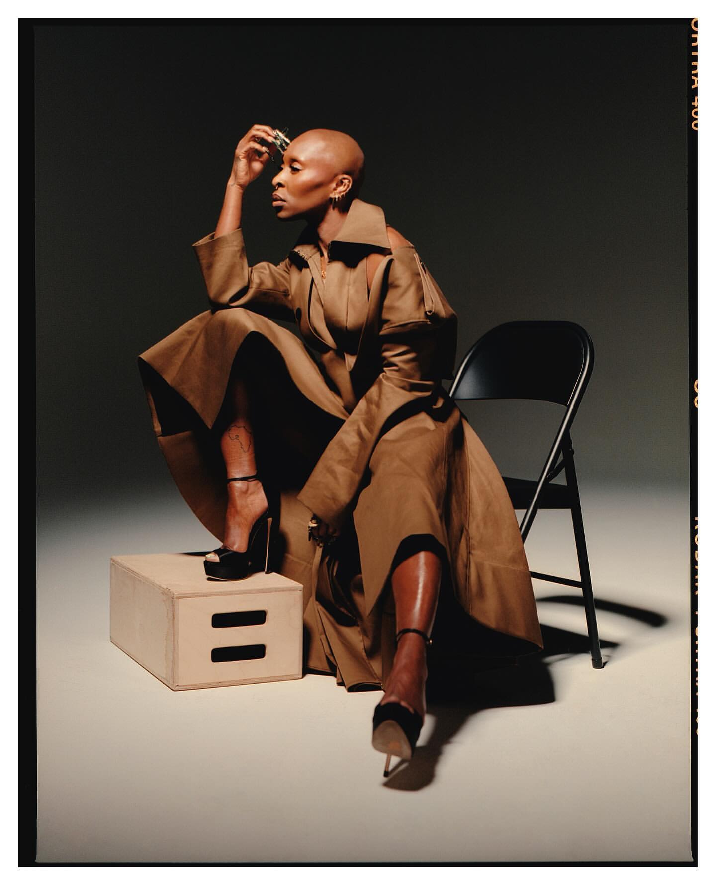 Elegant Black woman with shaved head in a brown trench coat and high heels poses seated on a wooden cube in a photo studio.