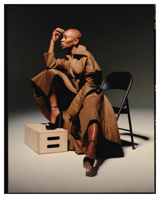 Elegant Black woman with shaved head in a brown trench coat and high heels poses seated on a wooden cube in a photo studio.