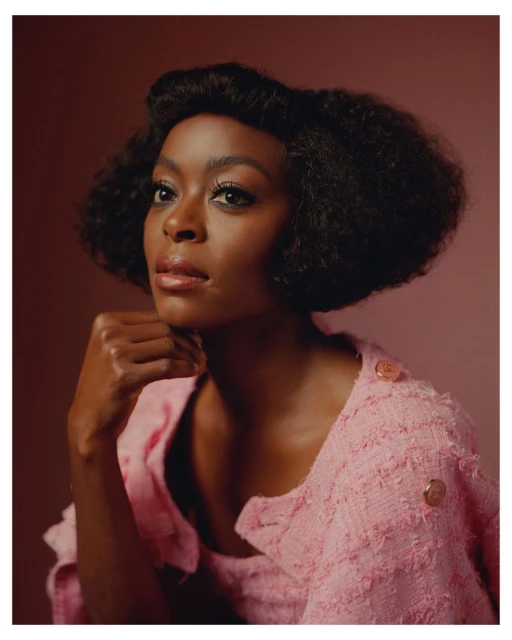 Portrait of a woman with dark skin and afro-textured hair wearing a pink jacket. She has her hand near her chin, looking upward.