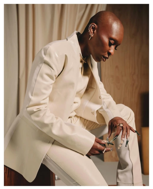 Bald adult in a white suit sitting on a wooden stool in front of a beige curtain; wearing jewelry and a neutral-toned suit.