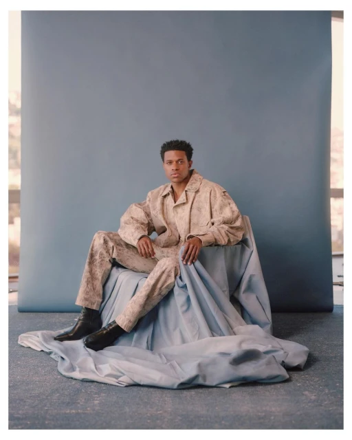 Young adult male poses in patterned beige suit and black boots against a blue backdrop, seated on draped fabric in a studio setting.