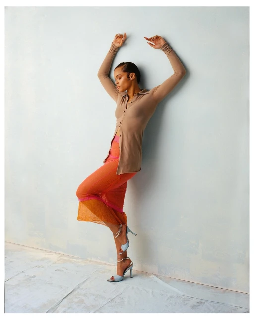 An adult female in a beige cardigan, sheer orange pants, and light blue high heels poses against a textured light blue wall in a studio.