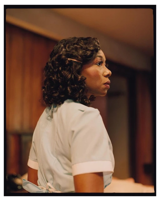 Woman with dark, curly hair in a light-blue uniform turns to the left with a neutral expression, set against a wood-panelled wall.