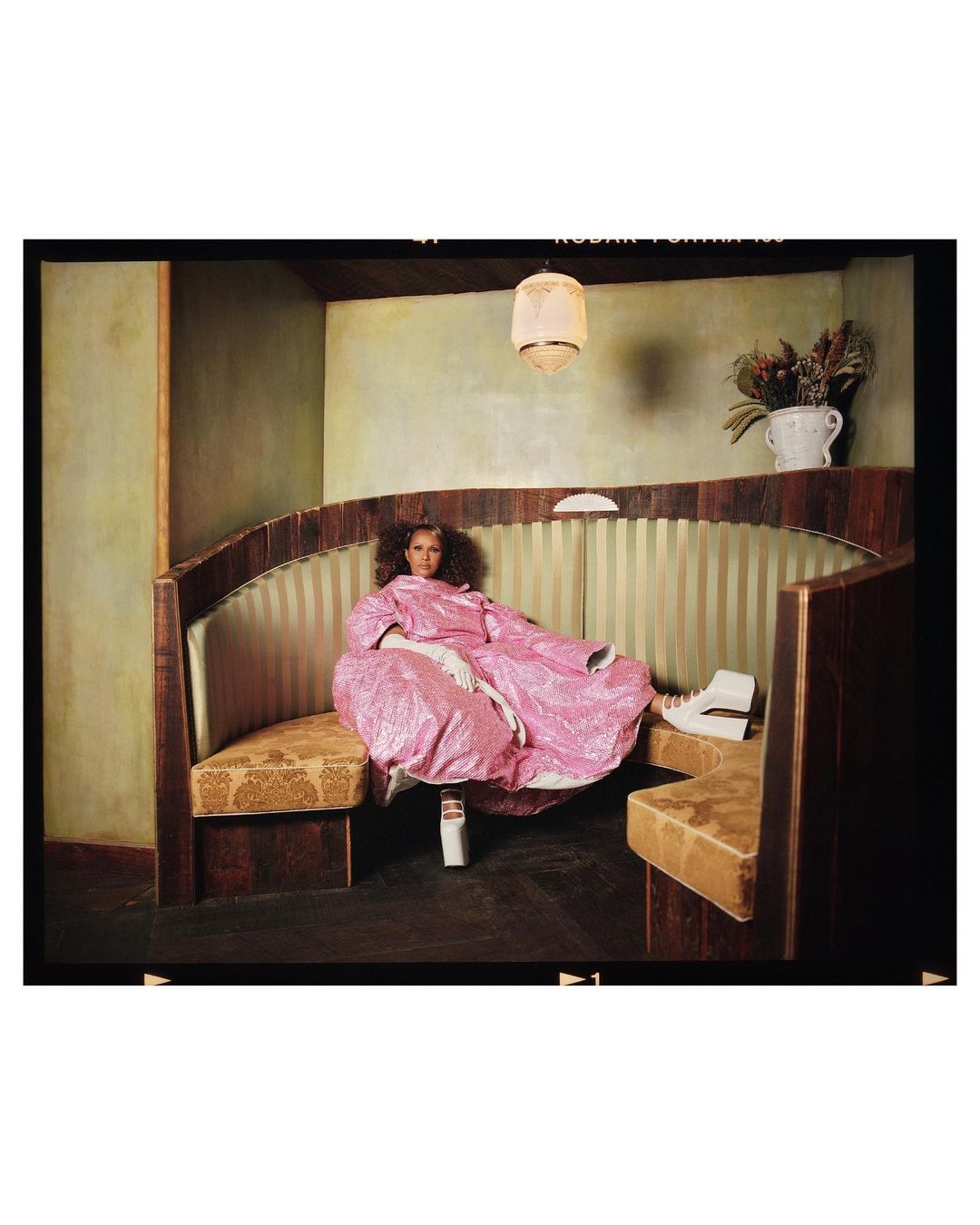 Woman in a pink robe poses in a vintage-style diner booth with white platform shoes, creating a unique fashion-forward image.