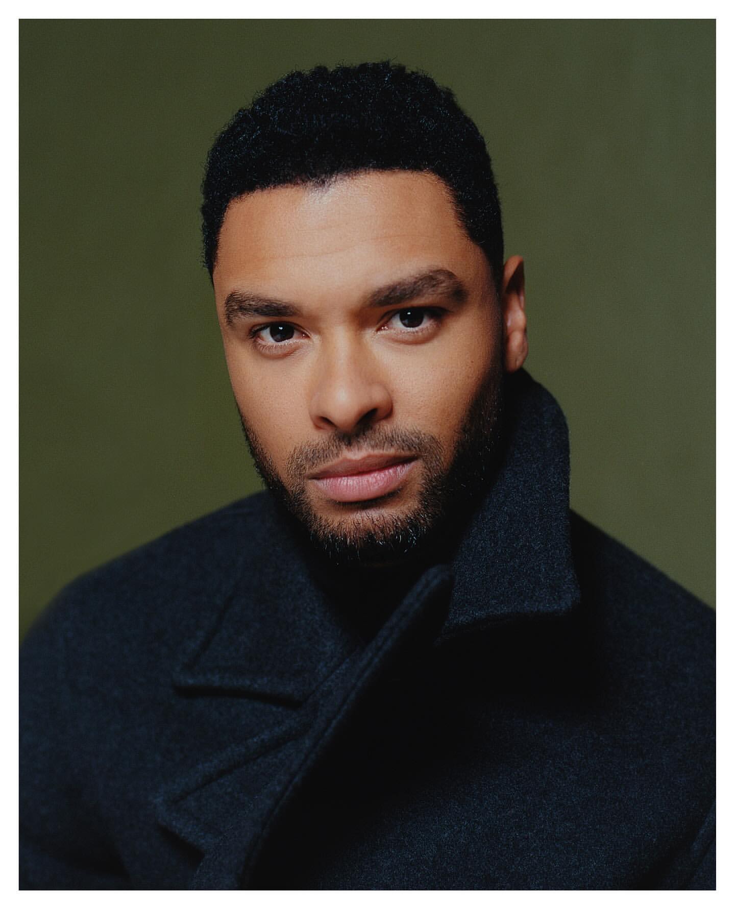 Close-up portrait of a handsome Black man in a dark coat with short, curly hair against an olive-green background, looking at the camera.