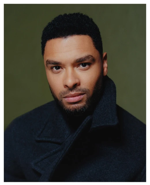 Close-up portrait of a handsome Black man in a dark coat with short, curly hair against an olive-green background, looking at the camera.
