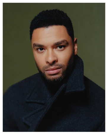 Close-up portrait of a handsome Black man in a dark coat with short, curly hair against an olive-green background, looking at the camera.