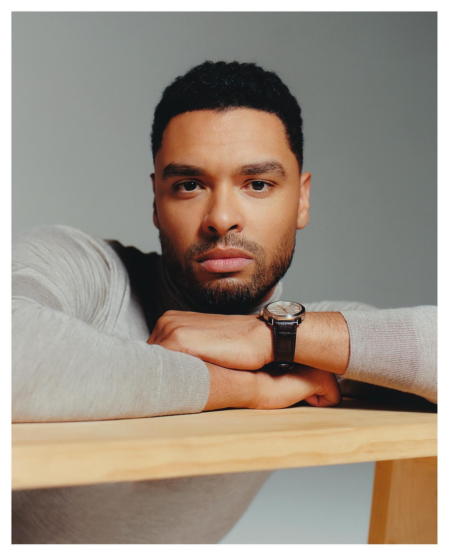 Portrait of a handsome adult male wearing a light grey turtleneck sweater and a wristwatch, resting his arms on a light wood table.