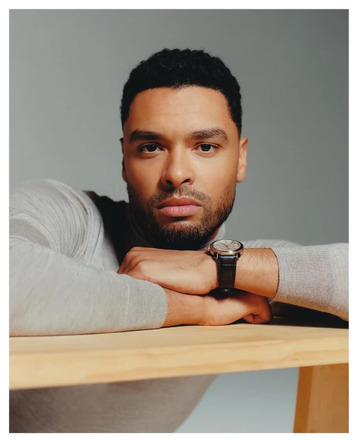 Portrait of a handsome adult male wearing a light grey turtleneck sweater and a wristwatch, resting his arms on a light wood table.