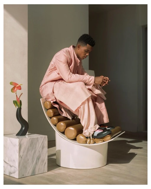 An adult male sits on a sculptural chair wearing a pink linen outfit indoors with a vase of flowers and a marble cube.