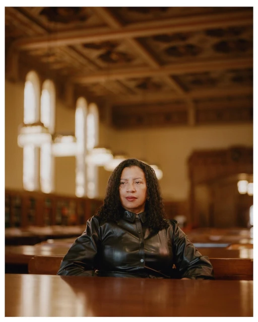 Portrait of a Black woman with curly hair in a leather jacket seated at a table, set in an ornate indoor library-style environment.