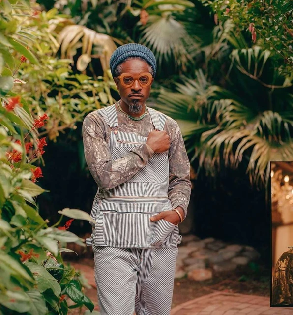 An adult man in striped overalls and glasses stands outdoors surrounded by lush greenery, foliage, and bright red flowers.
