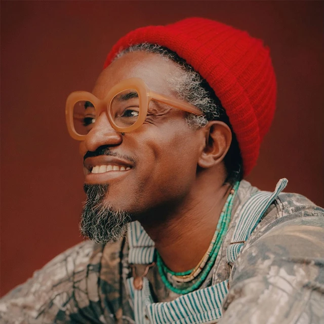 Portrait of smiling Black man with grey beard and red beanie, wearing statement glasses and layered necklaces against a brown backdrop.