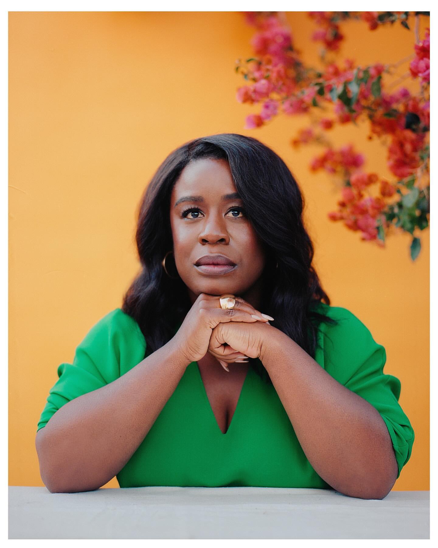 Portrait of a Black woman in a green dress with an orange background and pink flowers looking up with her hands clasped together.