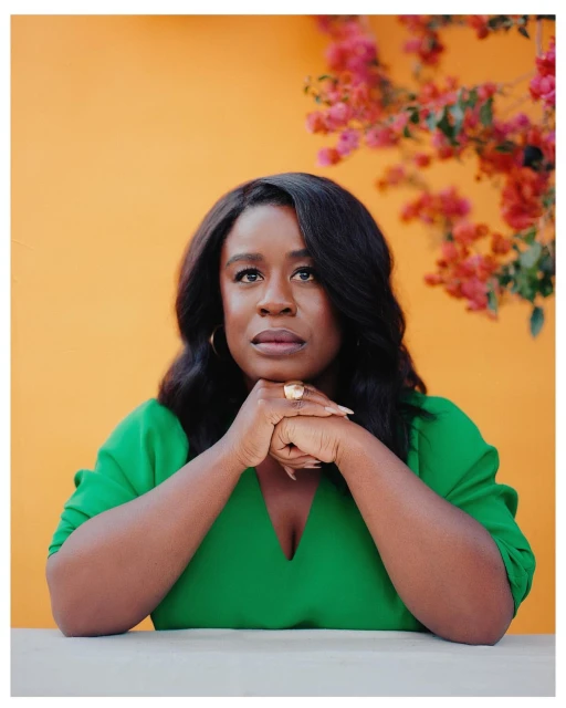 Portrait of a Black woman in a green dress with an orange background and pink flowers looking up with her hands clasped together.