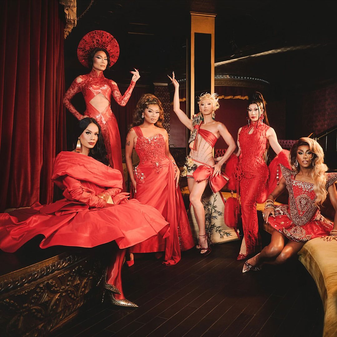 Group of six drag queens in elaborate red dresses pose indoors against a dark red curtain, showing off intricate details and vibrant colors.