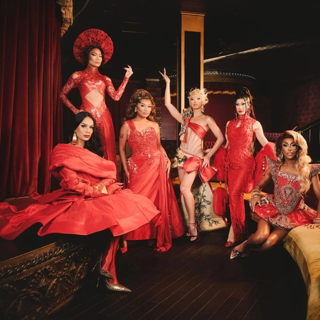 Group of six drag queens in elaborate red dresses pose indoors against a dark red curtain, showing off intricate details and vibrant colors.