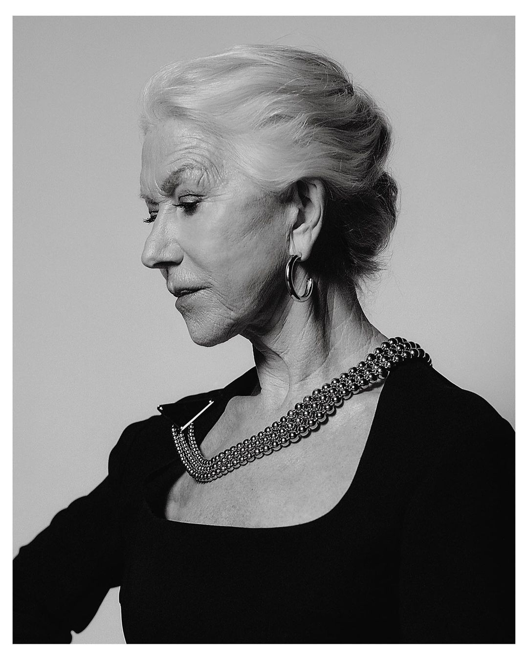 Monochrome portrait of Helen Mirren in profile wearing a dark top with a metallic beaded statement necklace and hoop earrings.
