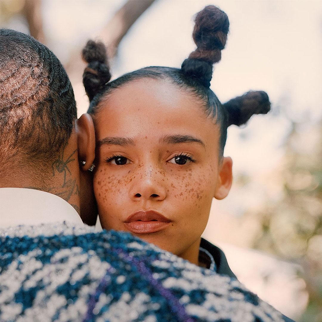 Portrait of an adult woman with an Afro hairstyle, embracing an adult male with neck tattoos in an outdoor setting with soft, diffused light.