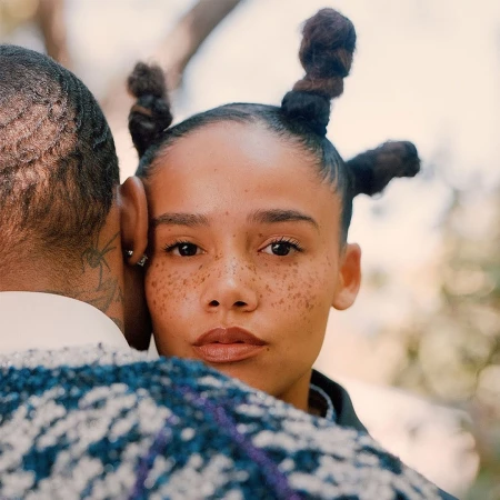 Portrait of an adult woman with an Afro hairstyle, embracing an adult male with neck tattoos in an outdoor setting with soft, diffused light.
