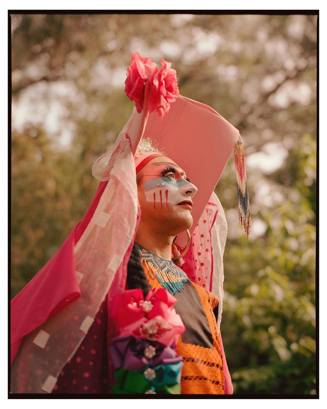 Person in colorful clothing with face paint and a headdress poses outdoors in a portrait orientation under natural light. 