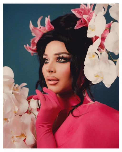 Close-up of a person with dramatic makeup, adorned with pink and white flowers, wearing a pink dress and matching gloves against a blue backdrop.