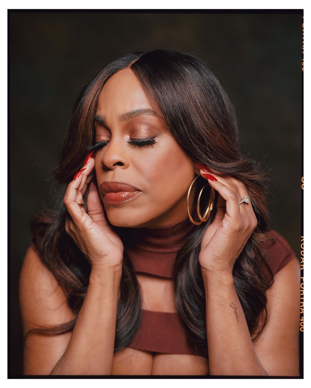 Portrait of African-American woman with auburn hair, wearing gold hoop earrings, against a dark brown background in a photo studio.