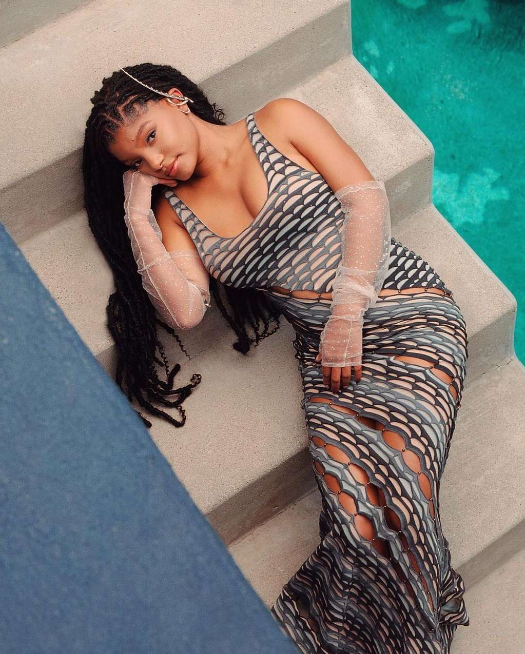 An adult woman with braided dark hair is lying on concrete steps near water, wearing a patterned gray dress with long sheer sleeves.