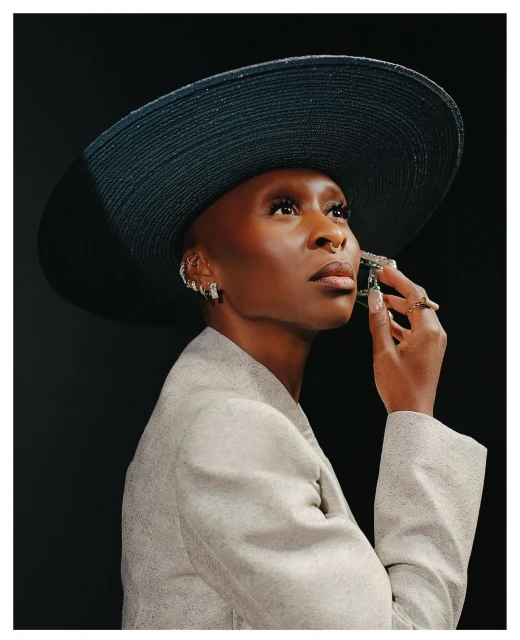Elegant portrait of a Black woman wearing a wide-brimmed hat and a neutral blazer against a dark backdrop, looking upwards.