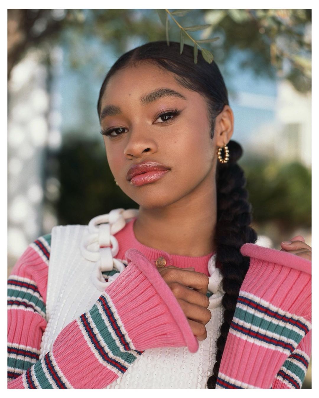 Portrait of a young woman with a braided ponytail wearing a pink-striped sweater and white top posing outdoors with leafy background.