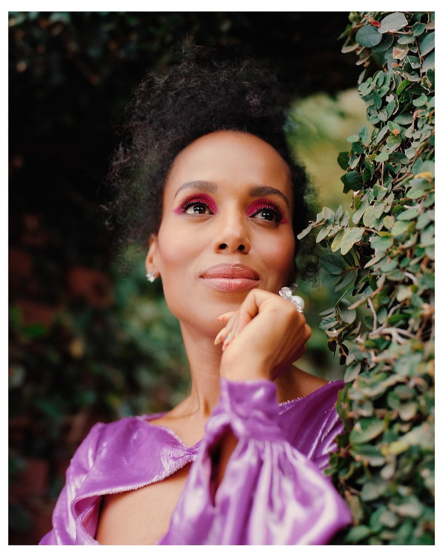 Close-up of a woman with curly hair and bright pink eyeshadow, posed in front of green foliage. She is wearing a shiny purple blouse.