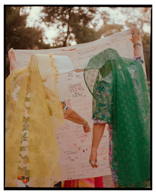 Two individuals hold a large, handwritten banner outdoors, partially obscured by patterned veils in yellow and green, trees in the background.
