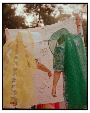 Two individuals hold a large, handwritten banner outdoors, partially obscured by patterned veils in yellow and green, trees in the background.