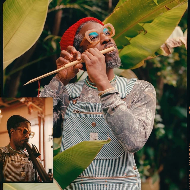 An adult Black man with a gray beard wearing overalls, glasses, and a red beanie is playing a flute while surrounded by green plants.