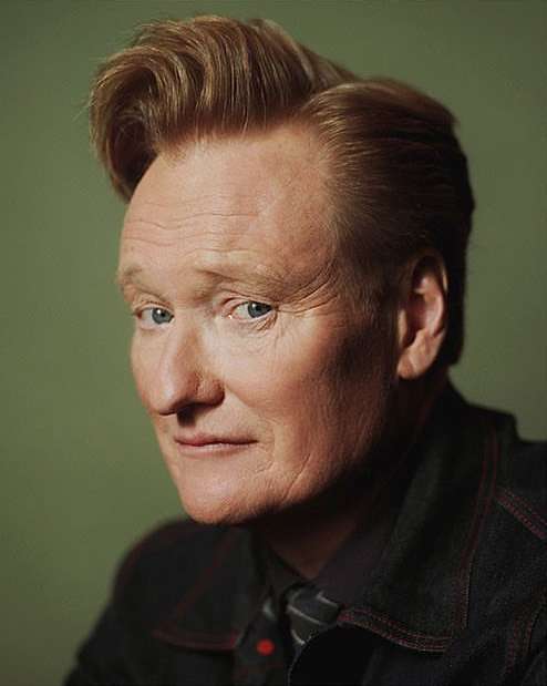 Close-up studio portrait of Conan O'Brien with auburn pompadour hairstyle, wearing a dark denim jacket and patterned necktie on light green background.