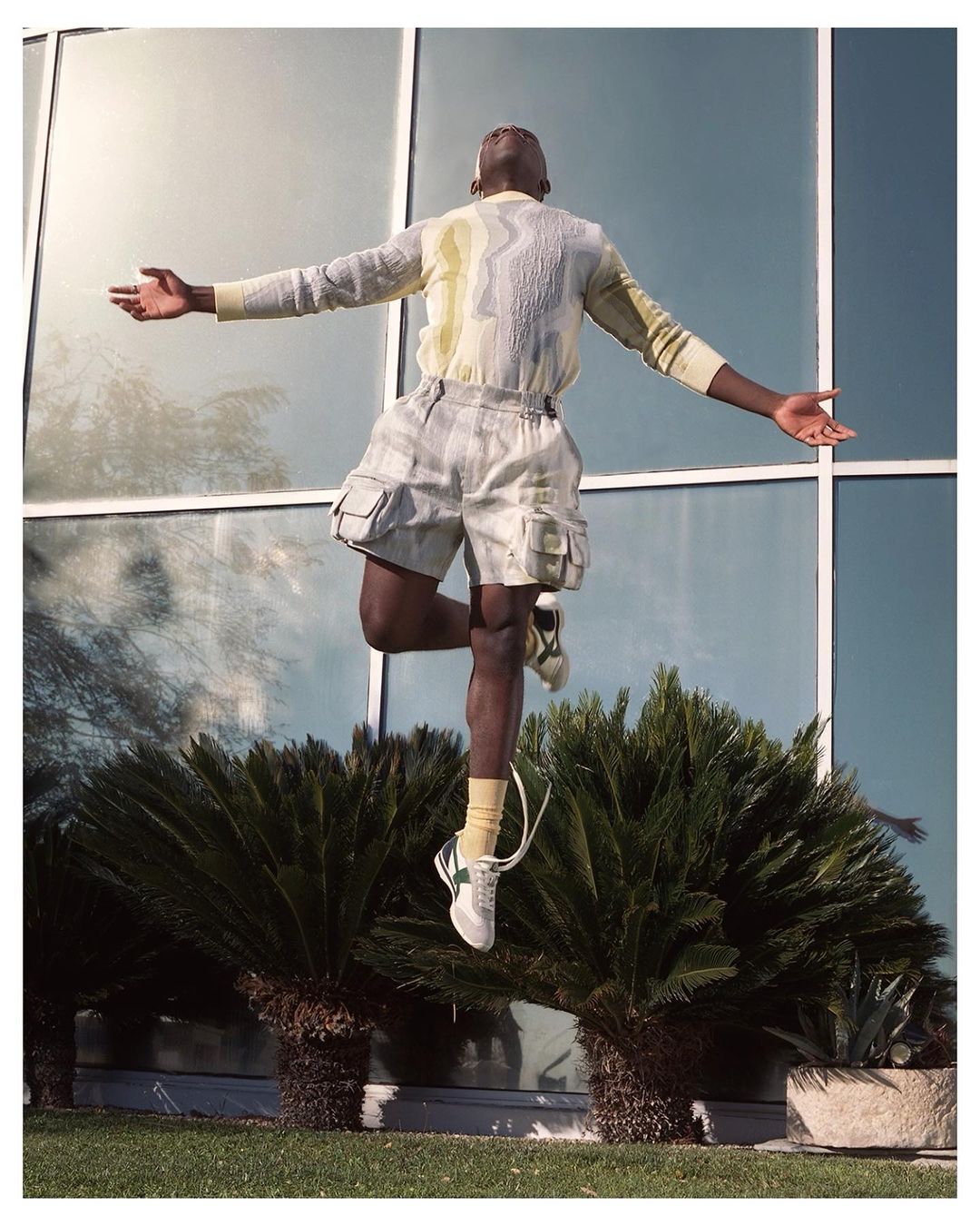 Athletic man leaps with arms outstretched in front of modern architecture and foliage. He wears a linen outfit and white sneakers.