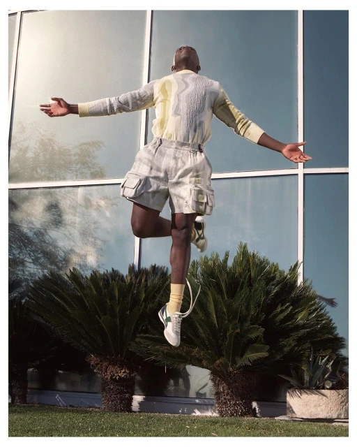 Athletic man leaps with arms outstretched in front of modern architecture and foliage. He wears a linen outfit and white sneakers.