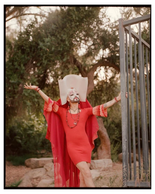 An adult male drag queen with face paint, red dress, and white hat is striking a pose in an outdoor park setting by a metal gate.
