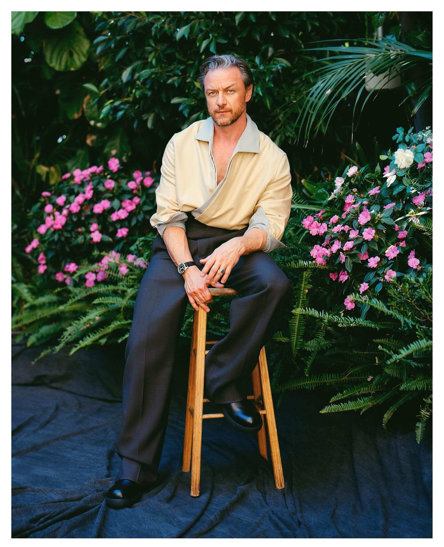 James McAvoy sits on a wooden stool in a collared shirt and dark pants, photographed outdoors among flowers and foliage.