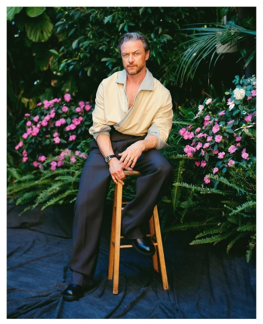 James McAvoy sits on a wooden stool in a collared shirt and dark pants, photographed outdoors among flowers and foliage.