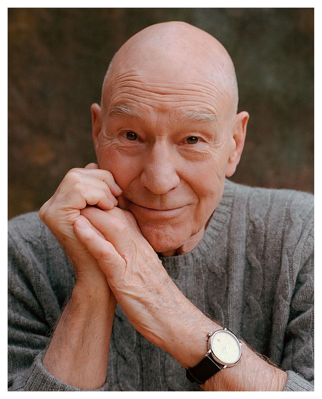 Portrait of Sir Patrick Stewart with hands clasped near his face, wearing a grey sweater and a wristwatch against a blurred background.