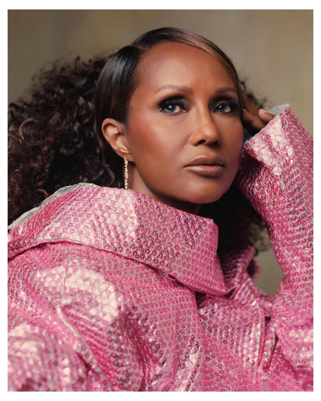Close-up portrait of a dark-skinned woman with brown curly hair and a shiny pink top looking directly at the camera in a studio setting.