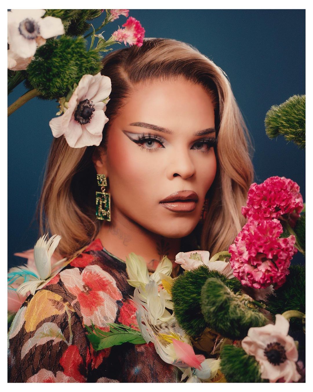 Portrait of an adult female with artistic makeup, floral dress, and statement earrings, surrounded by assorted cut flowers against a blue background.