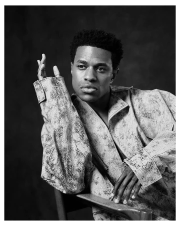 Black man in a textured jacket sitting on a chair against a dark background, photographed by Erik Carter in New York.