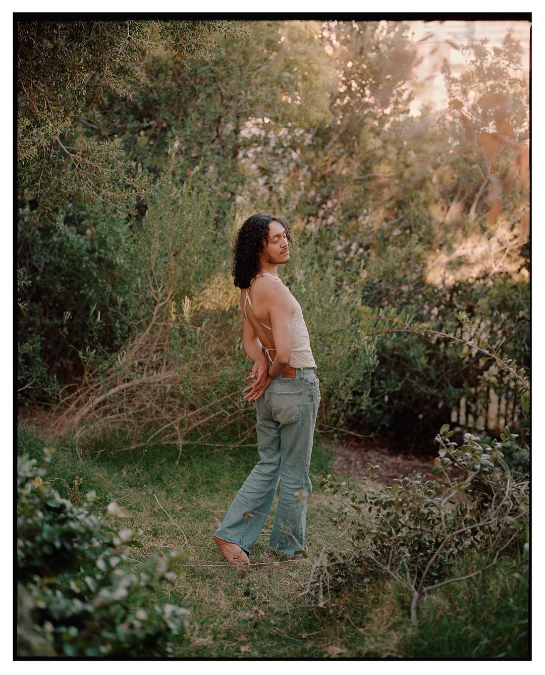 Person with long dark curly hair wearing light trousers and a backless top standing barefoot in a lush green garden.