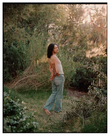 Person with long dark curly hair wearing light trousers and a backless top standing barefoot in a lush green garden.